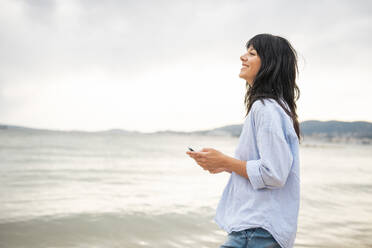 Lächelnde Frau mit langen Haaren hält Smartphone am Strand - JOSEF19230