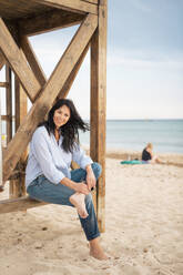 Smiling woman sitting on lifeguard hut at beach - JOSEF19248