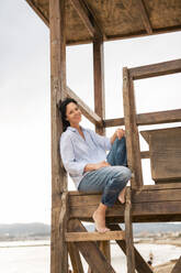 Mature smiling woman sitting on lifeguard hut at beach - JOSEF19260