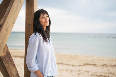 Smiling woman with hand in pocket leaning on lifeguard hut at beach - JOSEF19265