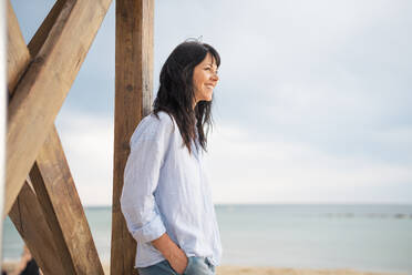 Happy mature woman with hand in pocket near lifeguard hut at beach - JOSEF19267