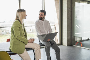 Businesswoman discussing with coworker holding laptop in office - UUF28677