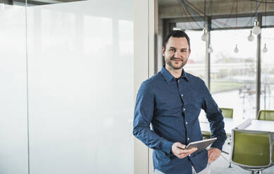 Selbstbewusster Geschäftsmann mit Tablet-PC an einer Glastür am Arbeitsplatz - UUF28682