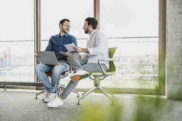 Business colleagues discussing over laptop and document sitting by glass window - UUF28699