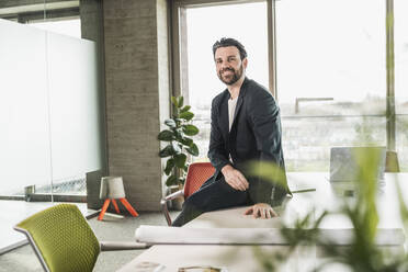 Confident businessman sitting on table in office - UUF28719