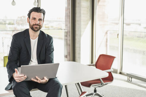 Selbstbewusster Geschäftsmann sitzt mit Laptop auf dem Tisch im Büro - UUF28720