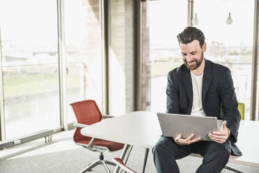 Smiling businessman sitting with laptop on table in office - UUF28721