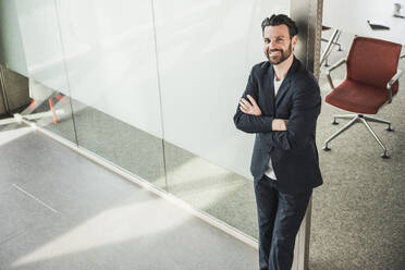 Confident businessman with arms crossed leaning on glass door - UUF28723