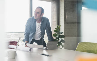 Thoughtful businessman leaning on table with paper and tablet PC in office - UUF28734