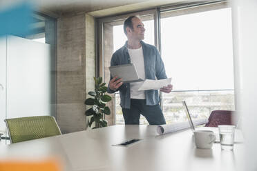 Thoughtful businessman holding paper and tablet PC in office - UUF28736