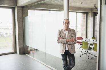 Confident businessman with arms crossed leaning on glass door in office - UUF28738