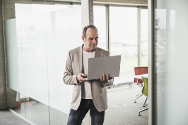 Businessman holding laptop standing by glass door in office - UUF28739