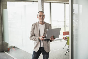 Confident businessman holding laptop standing by glass door in office - UUF28740