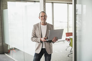 Selbstbewusster Geschäftsmann mit Laptop vor einer Glastür im Büro - UUF28740
