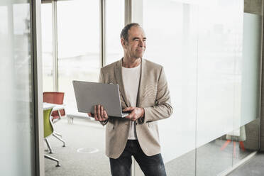 Smiling thoughtful businessman holding laptop standing by glass door in office - UUF28741