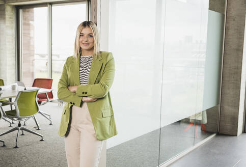 Confident young businesswoman with arms crossed standing by glass door in office - UUF28743