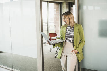 Smiling young businesswoman holding laptop standing by glass door in office - UUF28746