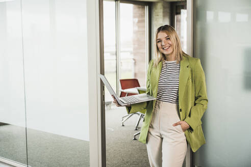 Selbstbewusste lächelnde Geschäftsfrau mit Laptop vor einer Glastür im Büro - UUF28747
