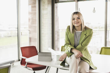 Confident smiling businesswoman sitting on table with tablet PC in office - UUF28748