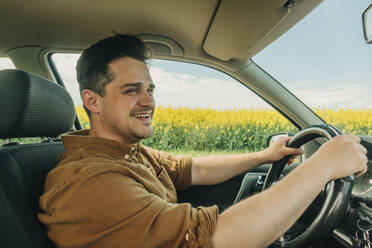 Smiling man driving car near rapeseed field - VSNF00892