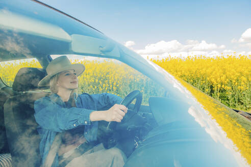 Lächelnde Frau am Steuer eines Autos in der Nähe eines Rapsfeldes, gesehen durch die Windschutzscheibe - VSNF00896