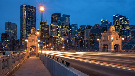 Long exposure of cityscape of Calgary city from pedestrian path with street lights and high rise contemporary buildings with glass walls reflecting colorful illumination under cloudless blue sky during dusk - ADSF44146