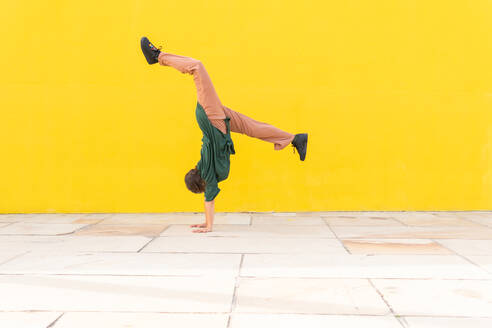 Ganzkörper-Seitenansicht einer anonymen flexiblen Frau in Freizeitkleidung, die einen Handstand auf einer gepflasterten Straße vor einer leuchtend gelben Wand macht - ADSF44153