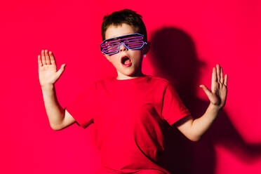 Excited boy wearing bright t shirt and stylish futuristic glasses standing on red background with opened mouth and raised arms - ADSF44166