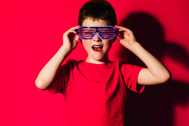 Aufgeregter Junge mit hellem T-Shirt und stilvoller futuristischer Brille, stehend auf rotem Hintergrund mit geöffnetem Mund - ADSF44167