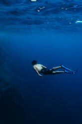 Ganzer Körper eines nicht erkennbaren Mannes mit Maske, Flossen und Badehose, der unter Wasser in einem blau kräuselnden Meer schwimmt - ADSF44168