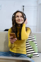 Positive young female in yellow turtleneck smiling and listening to music in headphones with smartphone looking up while sitting on sofa - ADSF44169