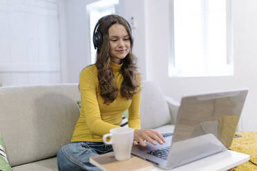 Concentrated young female freelancer in casual clothes sitting on sofa with cup of coffee in headphones browsing laptop while working remotely from home - ADSF44170