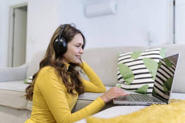 Side view of delighted young female freelancer in yellow turtleneck listening to music in wireless headphones looking at screen while working on laptop remotely from home - ADSF44171