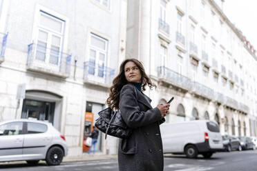 Seitenansicht einer selbstbewussten jungen Frau in einem warmen Mantel mit einer Tasche, die mit ihrem Handy telefoniert und wegschaut, während sie auf einer Straße in der Stadt steht - ADSF44175