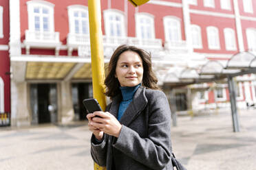 Positive junge Frau mit langen braunen Haaren in Mantel und blauem Rollkragenpullover steht auf der Straße in der Nähe eines roten Gebäudes und schreibt eine SMS mit dem Handy, während sie wegschaut - ADSF44176