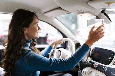 Seitenansicht einer positiven jungen Frau mit langen braunen gewellten Haaren in Freizeitkleidung, die Auto mit weißem Lenkrad fährt und dabei den Rückspiegel berührt - ADSF44183