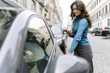 Smiling young woman with long brown wavy hair in casual clothes standing on paved street near modern car and opening door while holding smartphone - ADSF44190