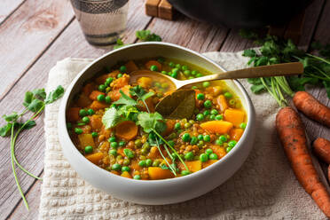 Von oben appetitlich frisch gekochte Suppe mit Karotten und grünen Erbsen, serviert in einer weißen Keramikschüssel auf einem Holztisch, dekoriert mit einem Petersilienzweig - ADSF44203