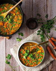 Draufsicht auf eine appetitliche, frisch gekochte Suppe mit Karotten und grünen Erbsen, serviert in einer weißen Keramikschüssel auf einem Holztisch, dekoriert mit einem Petersilienzweig - ADSF44204