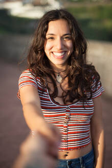 Glückliche junge Frau in Freizeitkleidung lächelt und schaut in die Kamera, während sie steht und die Hand ihres Freundes auf einem unscharfen Straßenhintergrund auf den Azoren in Portugal hält - ADSF44221
