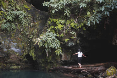 Rückenansicht einer nicht erkennbaren weiblichen Touristin, die mit ausgestreckten Händen auf einem umgestürzten Baumstamm in der Nähe eines Teiches und eines grünen Baumes in der Region der Azoren balanciert - ADSF44229