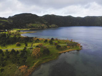 Luftaufnahme von ruhigen See in der Nähe von grünen grasbewachsenen Park mit Bäumen und Bergen unter bewölktem Himmel in Azoren - ADSF44235