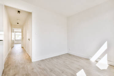 Interior design of spacious room with white walls and wooden floor in modern apartment with doorway leading to hallway in sunlight - ADSF44244