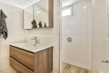 Inneneinrichtung eines modernen Duschbads mit weiß gefliesten Wänden und einem Fenster mit Sonnenschutzglas, während sich über dem Waschbecken ein Spiegelschrank mit Wasserhahn und Holzregalen befindet und Handtücher an Haltern hängen - ADSF44267