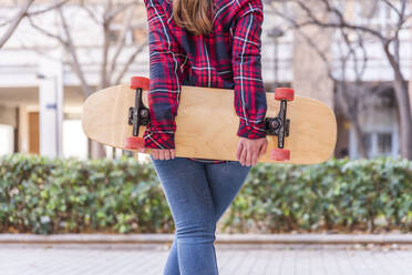 Rückenansicht einer anonymen Frau in Freizeitkleidung, die ein Skateboard hält, während sie auf dem Bürgersteig in einem Park steht - ADSF44306