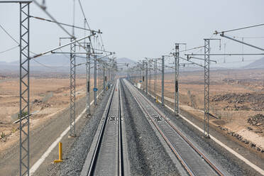 Luftaufnahme eines Infrastrukturentwicklungsprojekts mit der Verlegung von Eisenbahnschienen mit Oberleitungsmasten parallel zu Hochspannungsleitungen in trockenem Gelände während der Sommermonate - ADSF44324