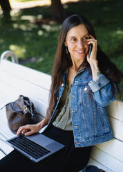 Ältere Frau mit Laptop und Smartphone sitzt auf einer Bank im Freien in einem Stadtpark und arbeitet. - HPIF15413