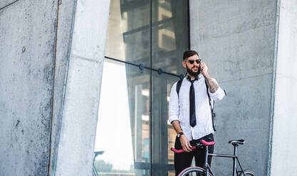 Portrait of young man commuter with bicycle standing outdoors in city, using smartphone. - HPIF15435