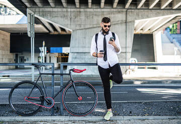 Porträt eines jungen Mannes, der mit seinem Fahrrad im Freien in der Stadt steht und sein Smartphone benutzt. - HPIF15437