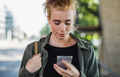 Front view portrait of young woman with red hair outdoors in town, using smartphone. - HPIF15447